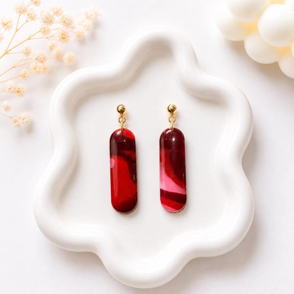 A pair of elongated pill-shaped dangle earrings with red and burgundy marble swirl pattern, hanging from small gold ball studs, displayed on a white wavy tray.