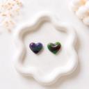 Two small heart-shaped clay stud earrings with multicolor marble patterns in purple and green tones, displayed on a white wavy ceramic tray.