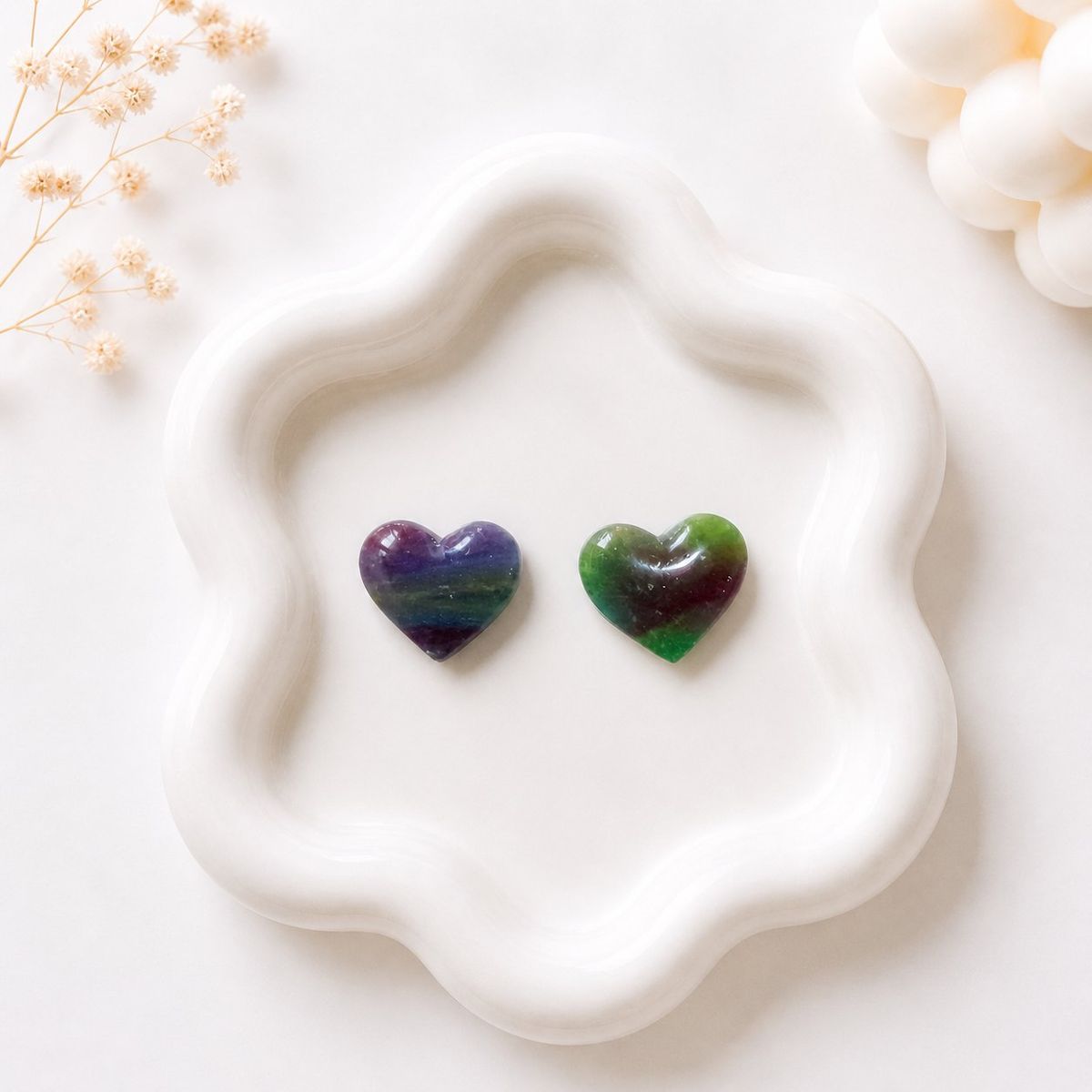 Two small heart-shaped clay stud earrings with multicolor marble patterns in purple and green tones, displayed on a white wavy ceramic tray.