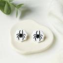 A pair of white polymer clay stud earrings shaped like spiderwebs with black spider details, displayed on a small cream ceramic dish.