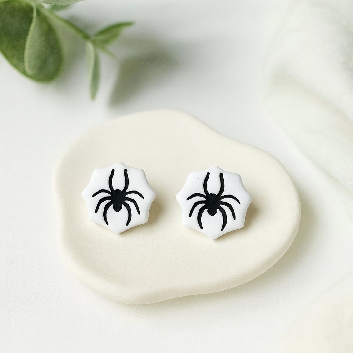 A pair of white polymer clay stud earrings shaped like spiderwebs with black spider details, displayed on a small cream ceramic dish.
