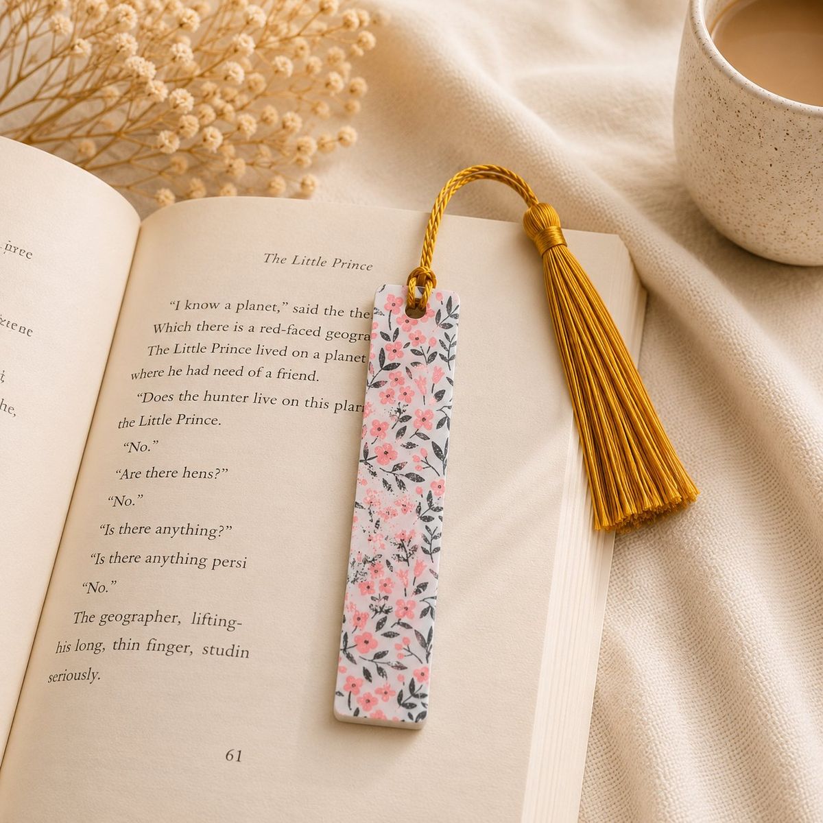 A rectangular white bookmark printed with pink flowers and dark leaves, topped with a gold twisted cord and gold tassel, resting on an open book.