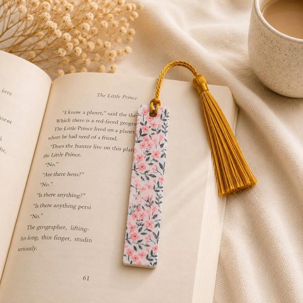 A rectangular white bookmark printed with pink flowers and dark leaves, topped with a gold twisted cord and gold tassel, resting on an open book.