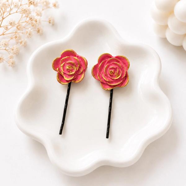 Two black bobby pins with pink and gold handmade clay rose toppers displayed on a white ceramic dish.