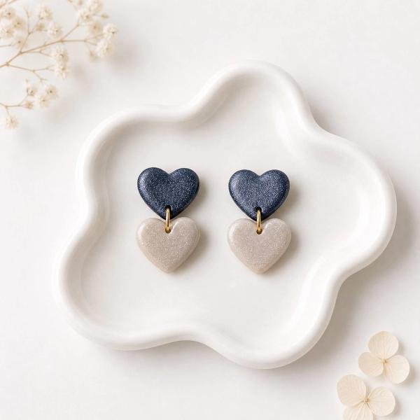A pair of handmade clay dangle earrings with a dark navy glitter heart stud and a beige shimmer heart drop connected by a small gold-tone ring, displayed on a white ceramic tray.