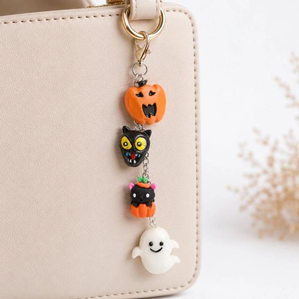 A Halloween bag charm with four polymer clay figures — an orange jack-o'-lantern, a black cat face, a black cat in a pumpkin, and a white ghost — hanging from a gold lobster clasp on a cream leather bag.