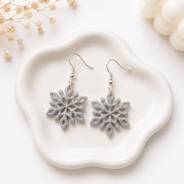 A pair of silver glitter snowflake dangle earrings with cutout detailing resting on a white ceramic dish.
