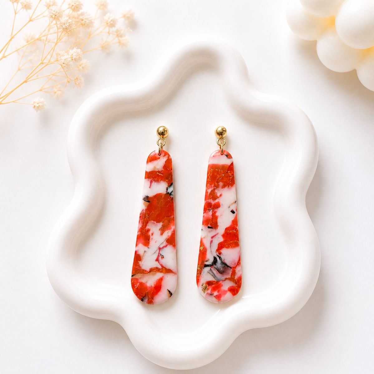 A pair of long tapered clay dangle earrings with a red, white, and black marble pattern and gold flecks, hanging from gold ball studs on a white ceramic tray.