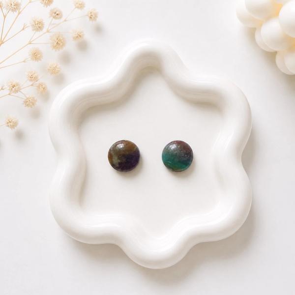 A pair of small round stud earrings with a marbled teal, purple, and brown pattern displayed on a white flower-shaped tray.