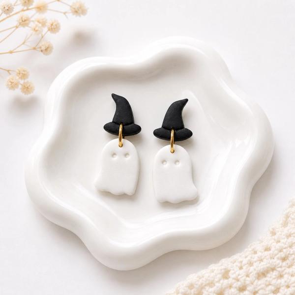 A pair of handmade clay dangle earrings with black witch hat studs and white ghost charms connected by gold-tone rings, displayed on a white ceramic dish.