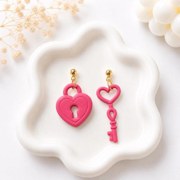A pair of mismatched dangle earrings on a white ceramic tray, one earring shaped as a hot pink clay heart padlock and the other as a hot pink clay heart-top key, both with gold ball studs.