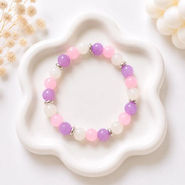 A stretch bracelet with alternating purple, pink, and white round beads separated by small silver disc spacers, displayed on a white flower-shaped ceramic tray.