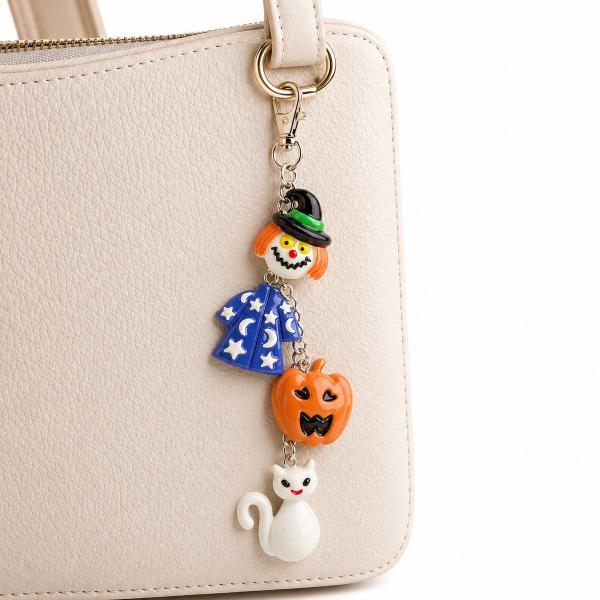 A Halloween-themed bag charm with four colorful resin figures — a clown, a blue star wizard cape, an orange jack-o'-lantern, and a white cat — linked on a silver chain with a gold lobster clasp, attached to a cream leather handbag.