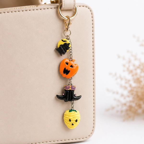 A Halloween-themed polymer clay key charm with four linked charms — a bat on a yellow moon, an orange jack-o'-lantern, a black witch hat, and a yellow mummy strawberry — attached to a beige handbag via a gold lobster clasp.