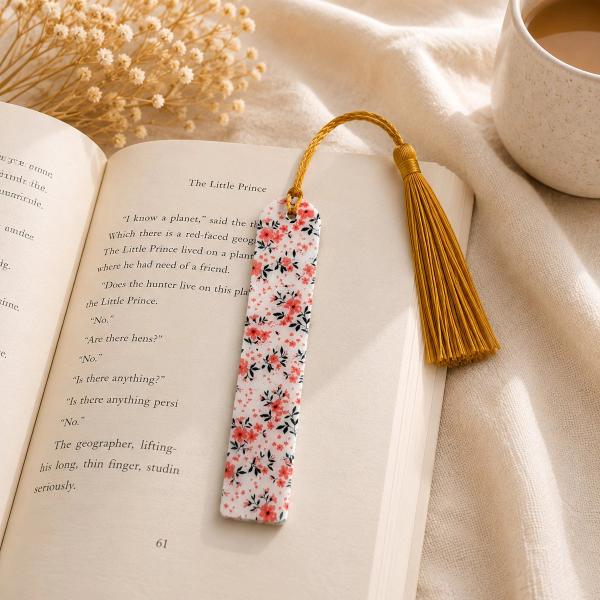 A rectangular white bookmark covered in a small pink floral and dark leaf print, with a gold tassel attached at the top, resting on an open book.