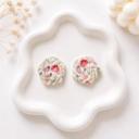 A pair of round white clay stud earrings with a twisted texture and red and green floral print, displayed on a white decorative tray.