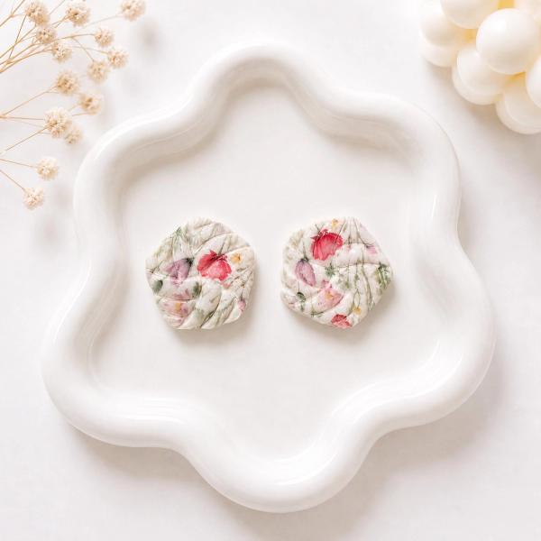 A pair of round white clay stud earrings with a twisted texture and red and green floral print, displayed on a white decorative tray.