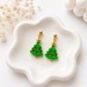 A pair of green clay Christmas tree dangle earrings with red berry details, gold star toppers, and gold ball stud posts resting in a white ceramic dish.