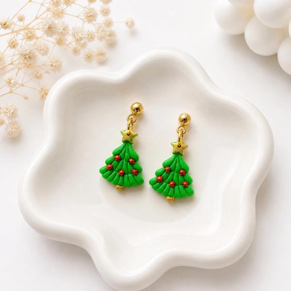 A pair of green clay Christmas tree dangle earrings with red berry details, gold star toppers, and gold ball stud posts resting in a white ceramic dish.