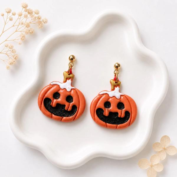 A pair of handmade orange clay jack-o-lantern dangle earrings with black carved faces, white drip detail, and gold ball stud posts displayed on a white ceramic tray.