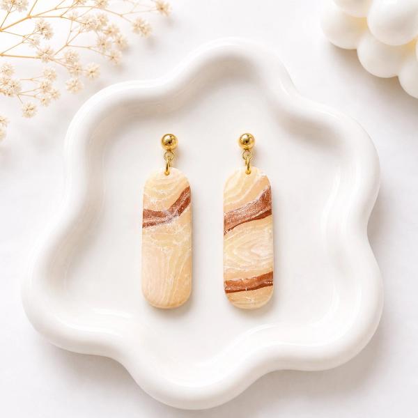 A pair of elongated rounded clay dangle earrings with a beige and brown marble swirl pattern, hung from gold ball studs, displayed on a white wavy ceramic tray.
