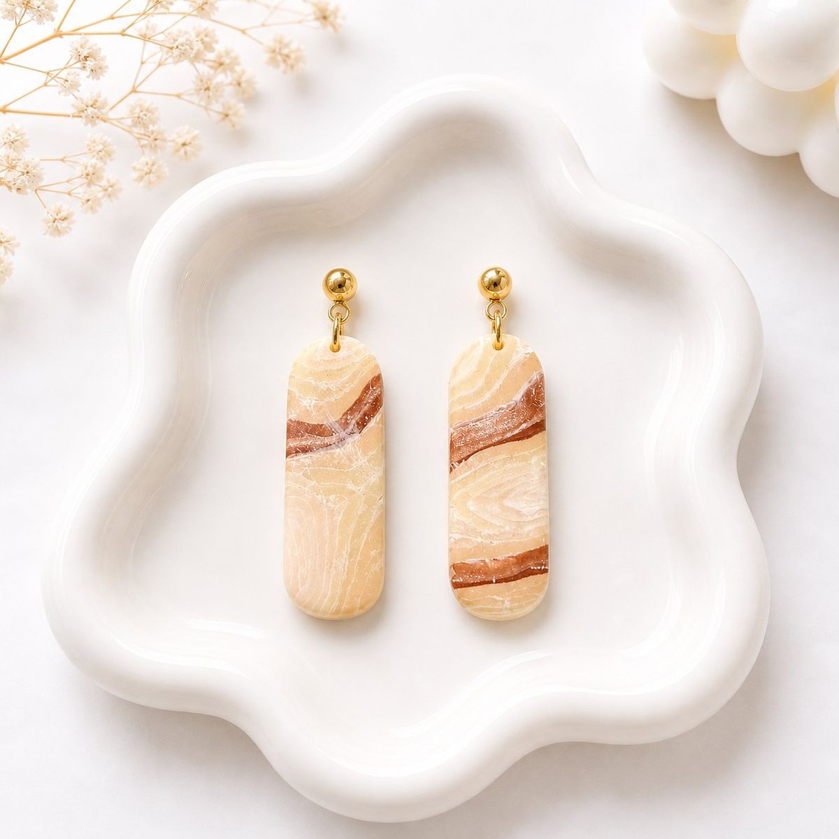 A pair of elongated rounded clay dangle earrings with a beige and brown marble swirl pattern, hung from gold ball studs, displayed on a white wavy ceramic tray.