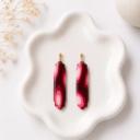 A pair of elongated clay dangle earrings with a burgundy and pink marble swirl pattern and scalloped edges, attached to gold ball studs, displayed on a white ceramic tray.