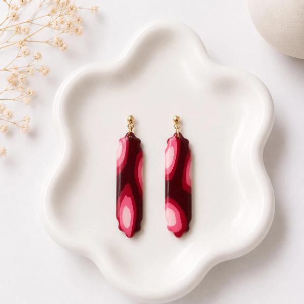 A pair of elongated clay dangle earrings with a burgundy and pink marble swirl pattern and scalloped edges, attached to gold ball studs, displayed on a white ceramic tray.