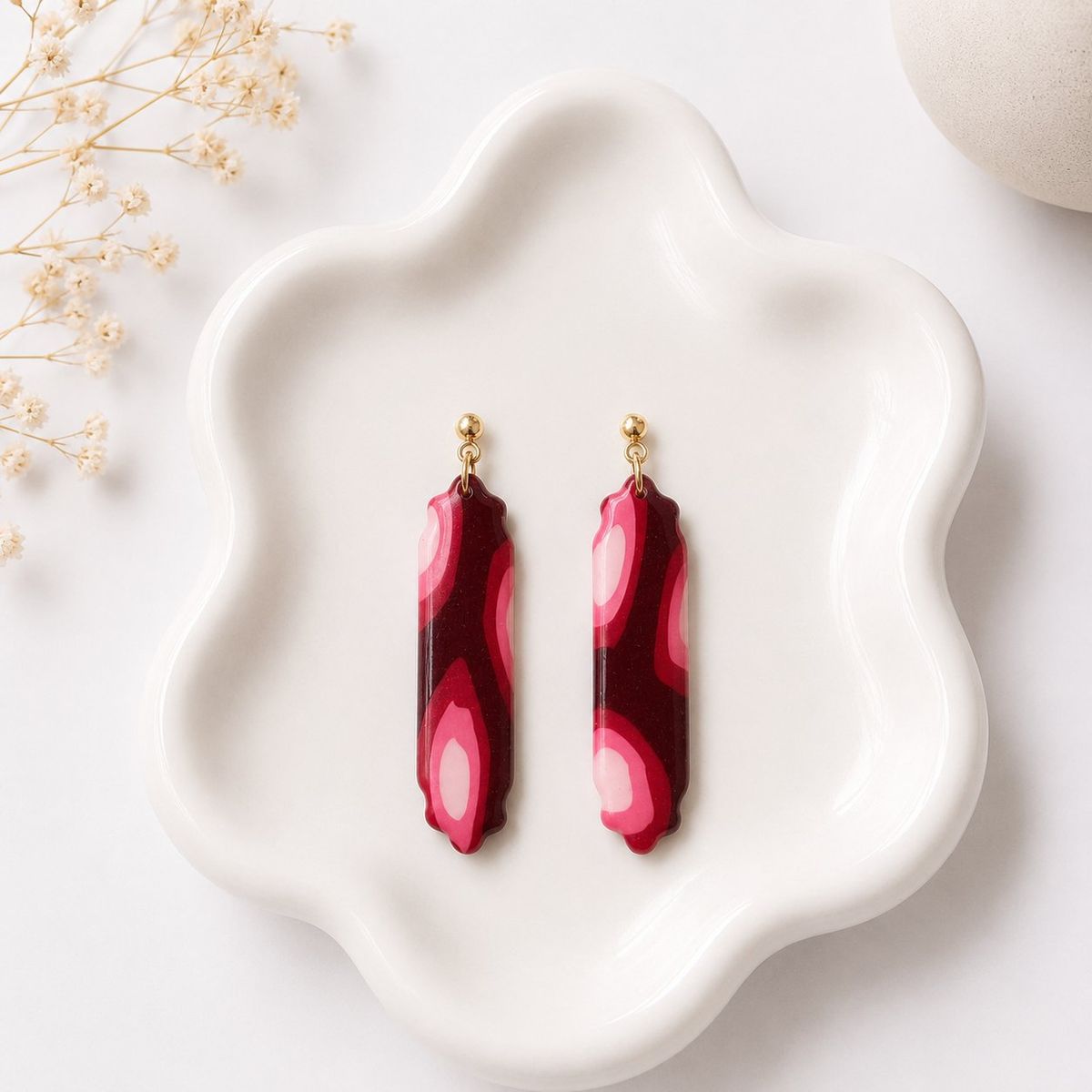 A pair of elongated clay dangle earrings with a burgundy and pink marble swirl pattern and scalloped edges, attached to gold ball studs, displayed on a white ceramic tray.