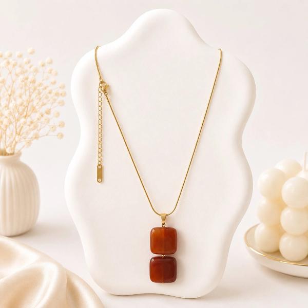 A gold snake chain necklace with two stacked square terracotta red stone pendants displayed on a white jewelry bust.