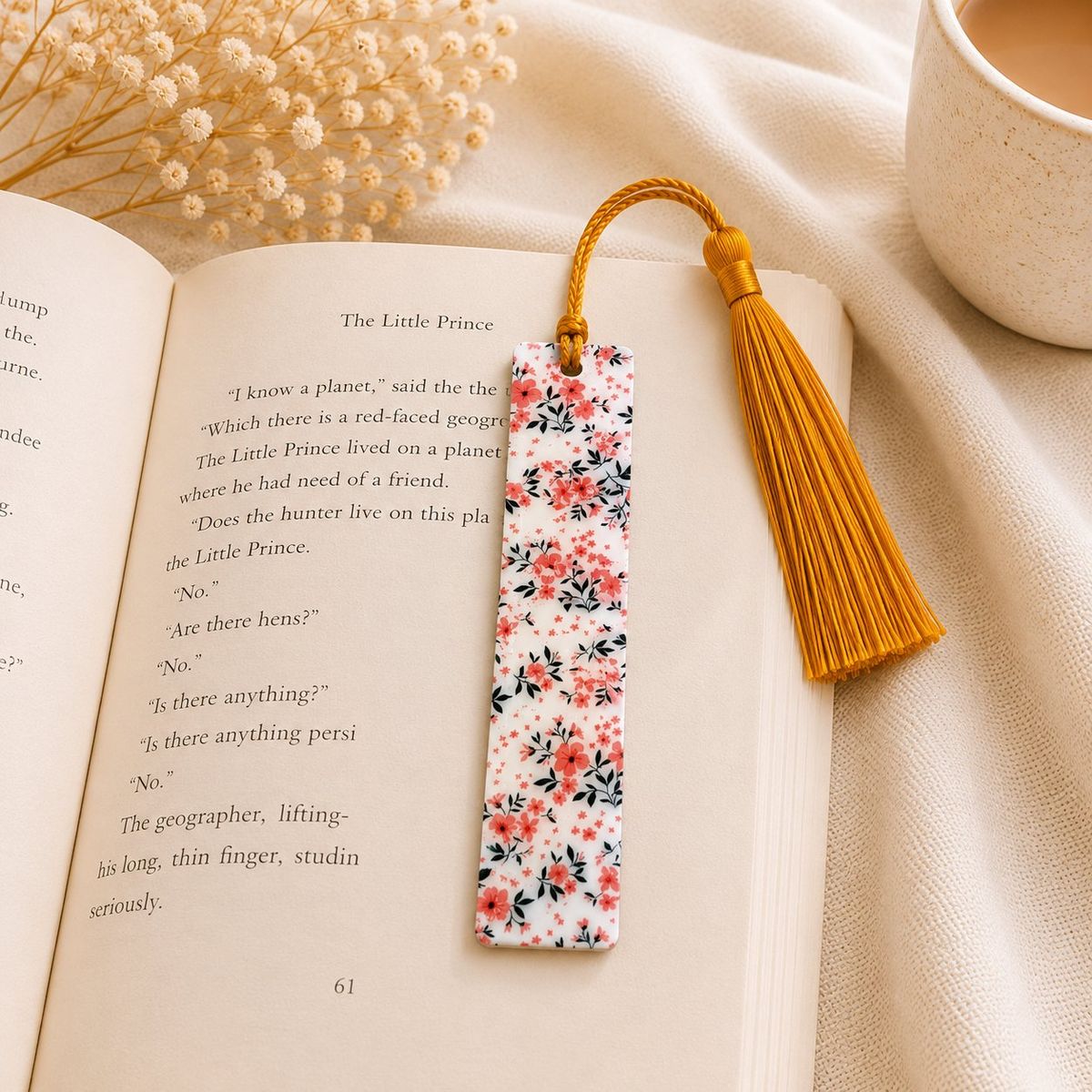 A rectangular white bookmark covered in a pink floral and black leaf pattern, with a mustard yellow tassel attached at the top, resting on an open book.