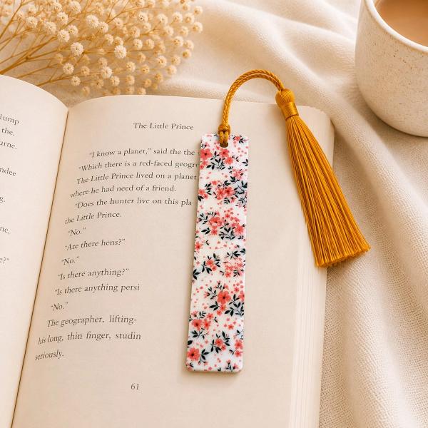 A rectangular white bookmark covered in a pink floral and black leaf pattern, with a mustard yellow tassel attached at the top, resting on an open book.