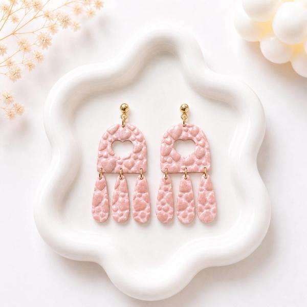 A pair of blush pink polymer clay dangle earrings with an embossed heart pattern, arch-shaped top with a heart cutout, and three hanging teardrop pieces on gold jump rings with gold ball studs.