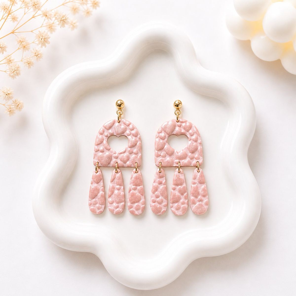 A pair of blush pink polymer clay dangle earrings with an embossed heart pattern, arch-shaped top with a heart cutout, and three hanging teardrop pieces on gold jump rings with gold ball studs.