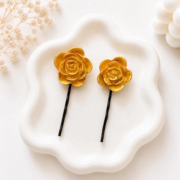 Two gold clay rose bobby pins of different sizes displayed on a white ceramic tray with dried flowers in the background.