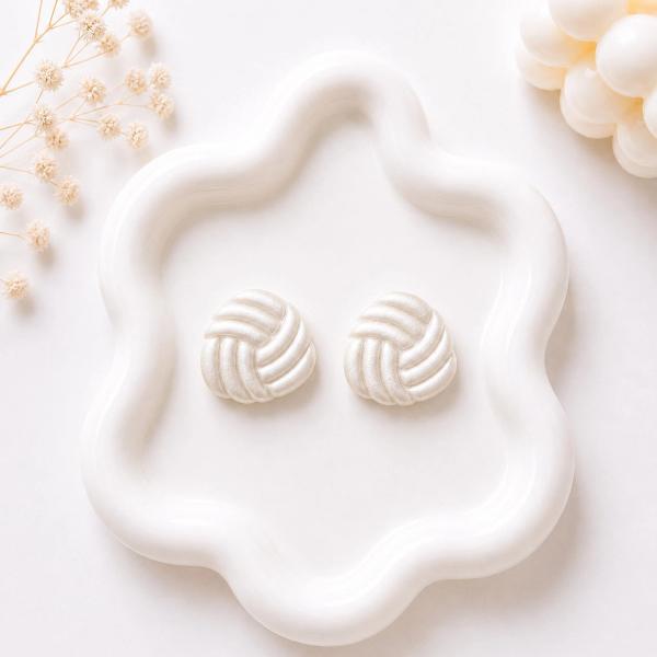A pair of round white pearlescent clay stud earrings with a woven knot texture, displayed on a white wavy tray.