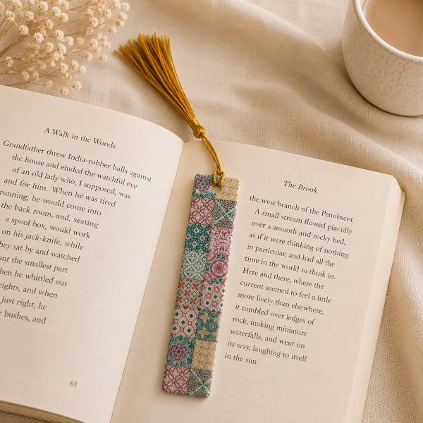 A rectangular bookmark with a colorful mosaic tile pattern in teal, pink, and gold, topped with a gold tassel, resting on an open book.