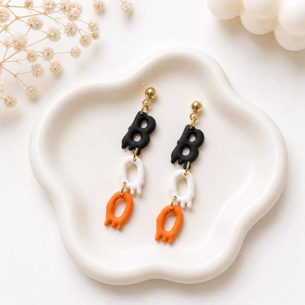 A pair of handmade clay dangle earrings with three linked charms spelling BOO in black, white, and orange, attached to gold ball studs, displayed on a white ceramic tray.