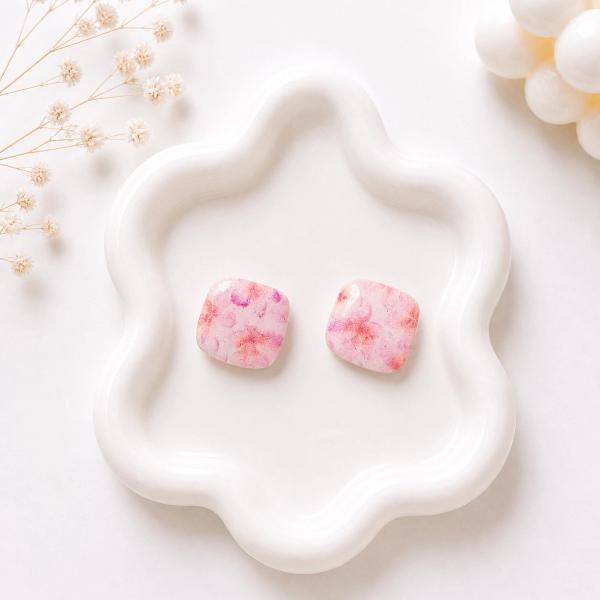 A pair of rounded square stud earrings with a pink and peach floral pattern displayed on a white wavy ceramic tray.