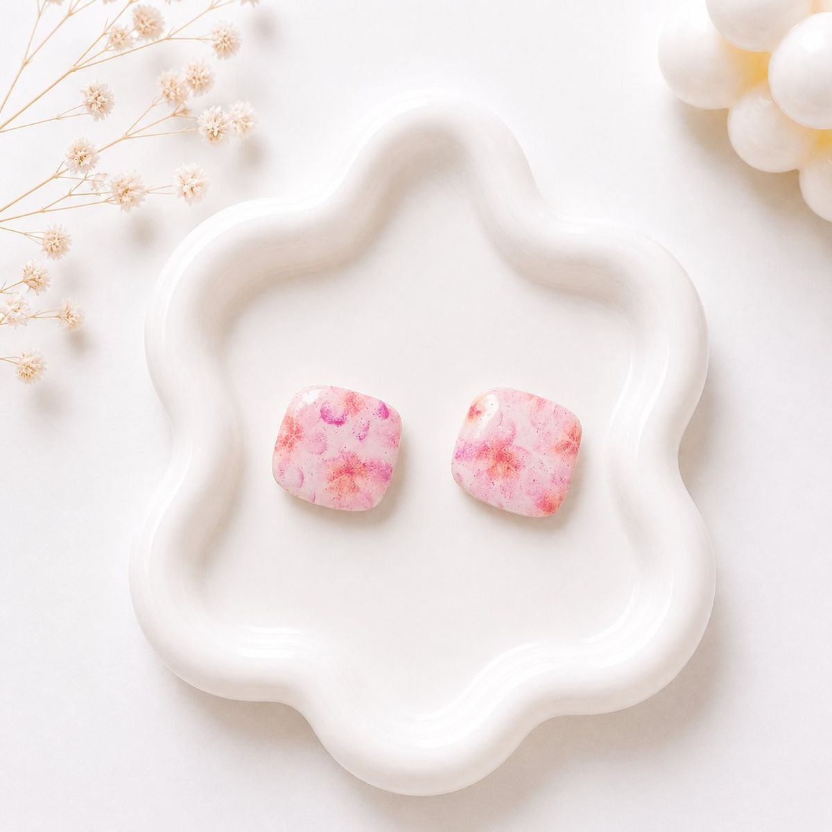 A pair of rounded square stud earrings with a pink and peach floral pattern displayed on a white wavy ceramic tray.