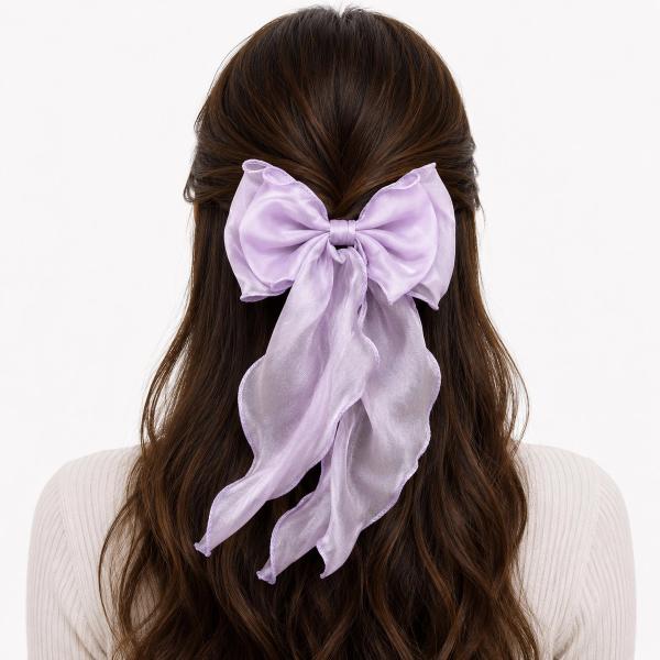 A large lavender sheer satin bow clip with scalloped edges and long flowing ribbon tails, worn in the half-up hair of a brunette woman.