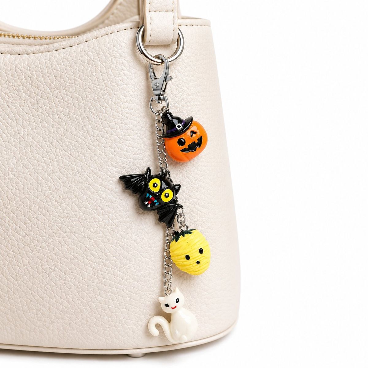 A silver-tone keychain with four Halloween resin charms — an orange jack-o'-lantern with witch hat, a black bat, a yellow pumpkin face, and a white ghost cat — clipped onto a cream leather handbag.