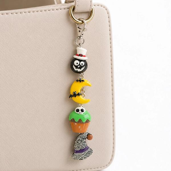 A handmade polymer clay bag charm with four linked Halloween motifs — a black jack-o'-lantern with a white top hat, a yellow crescent moon with bats, a green skull cupcake, and a spider web witch boot — attached to a beige leather handbag via a lobster clasp.