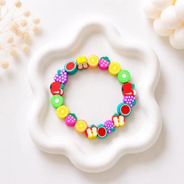 A circular stretch bracelet made of multicolored polymer clay fruit-shaped beads including watermelon, kiwi, orange, grape, strawberry, and apple, displayed on a white wavy ceramic dish.