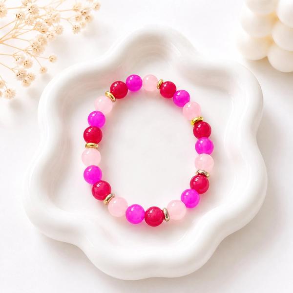 A stretch bracelet with round beads in blush pink, hot pink, and magenta, separated by small gold-tone disc spacers, displayed on a white ceramic tray.