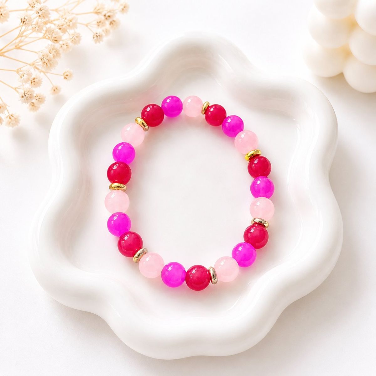 A stretch bracelet with round beads in blush pink, hot pink, and magenta, separated by small gold-tone disc spacers, displayed on a white ceramic tray.