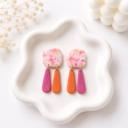 A pair of handmade clay dangle earrings with marbled pink and orange square tops and two teardrop-shaped charms in solid magenta and orange, displayed on a white ceramic tray.