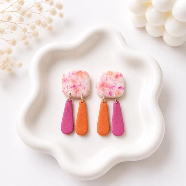 A pair of handmade clay dangle earrings with marbled pink and orange square tops and two teardrop-shaped charms in solid magenta and orange, displayed on a white ceramic tray.