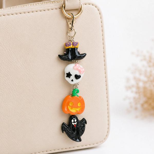 A Halloween key chain charm featuring four linked resin charms — a black witch hat, white skull with pink bow, orange jack-o'-lantern, and black ghost — clipped to a beige handbag via a gold lobster clasp.