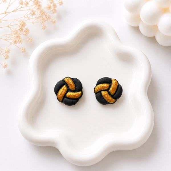 A pair of round black clay stud earrings with a braided knot pattern and gold-painted grooves, displayed on a white ceramic tray.